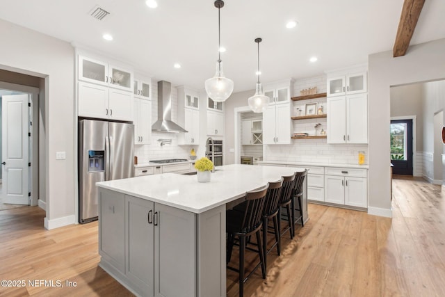 kitchen featuring beamed ceiling, white cabinets, stainless steel appliances, a center island with sink, and wall chimney exhaust hood