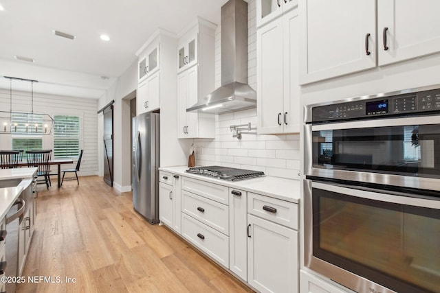 kitchen featuring pendant lighting, white cabinets, stainless steel appliances, wall chimney range hood, and light hardwood / wood-style flooring