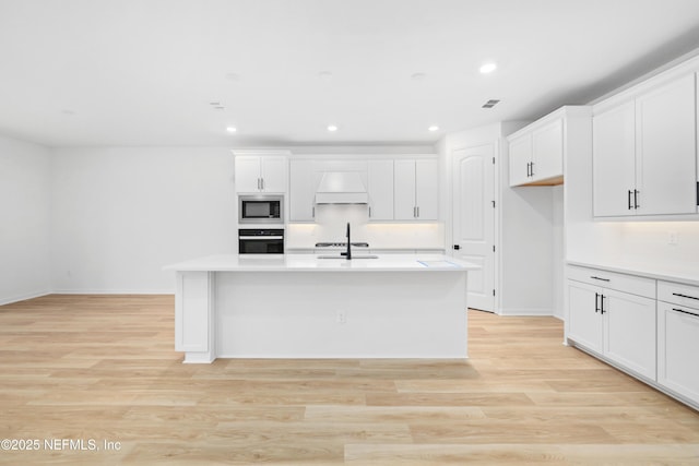 kitchen with black oven, light hardwood / wood-style flooring, premium range hood, stainless steel microwave, and a center island with sink