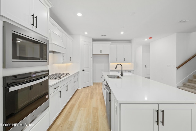 kitchen with sink, stainless steel appliances, an island with sink, white cabinets, and light wood-type flooring