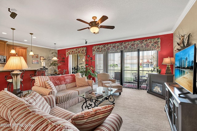 living room with a ceiling fan, a glass covered fireplace, light colored carpet, a textured ceiling, and crown molding