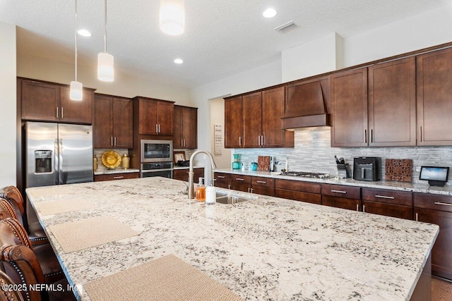 kitchen with premium range hood, sink, hanging light fixtures, appliances with stainless steel finishes, and an island with sink