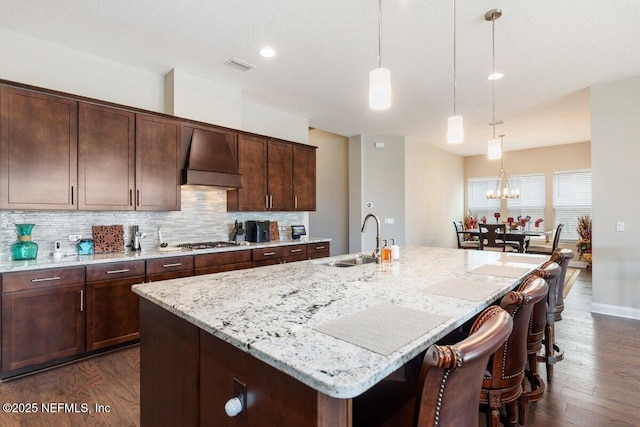 kitchen featuring sink, a kitchen island with sink, hanging light fixtures, stainless steel gas cooktop, and wall chimney exhaust hood