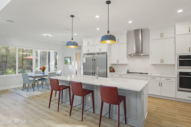 kitchen featuring wall chimney range hood, an island with sink, pendant lighting, stainless steel appliances, and white cabinets