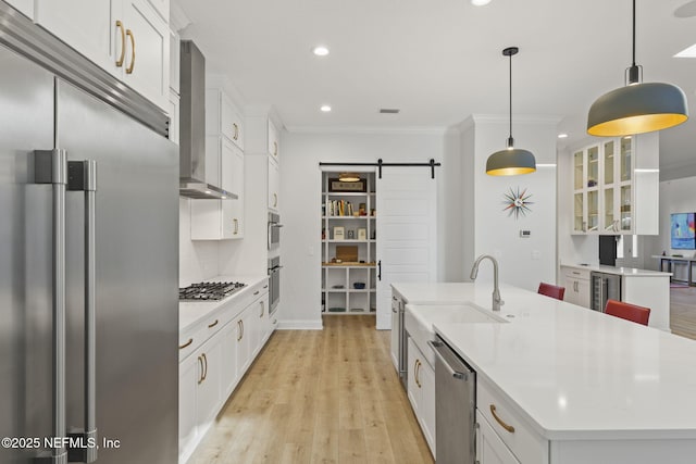 kitchen featuring white cabinets, a center island with sink, stainless steel appliances, a barn door, and wall chimney range hood