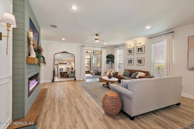 living room featuring crown molding, a textured ceiling, light wood-type flooring, a large fireplace, and ceiling fan