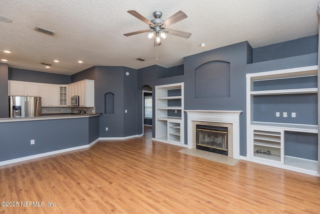 unfurnished living room featuring a textured ceiling, light wood-type flooring, built in features, ceiling fan, and a tiled fireplace