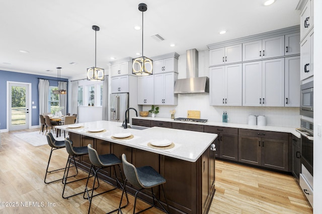 kitchen with an island with sink, a breakfast bar area, hanging light fixtures, stainless steel appliances, and wall chimney range hood