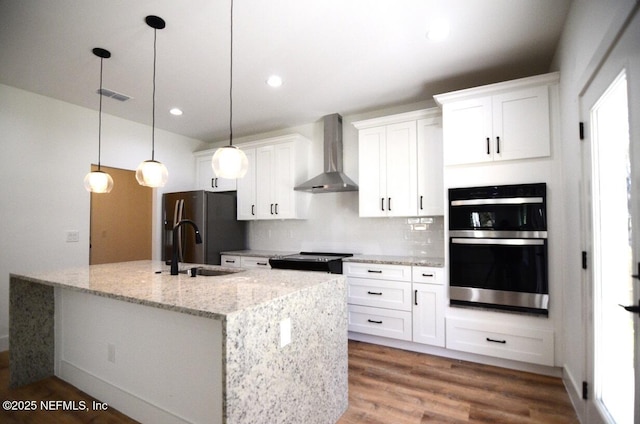 kitchen with pendant lighting, wall chimney exhaust hood, white cabinetry, an island with sink, and light stone counters