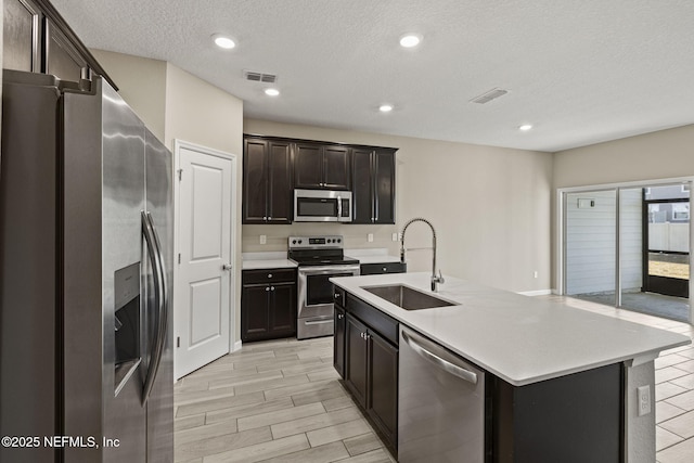 kitchen with sink, a kitchen island with sink, stainless steel appliances, dark brown cabinets, and a textured ceiling