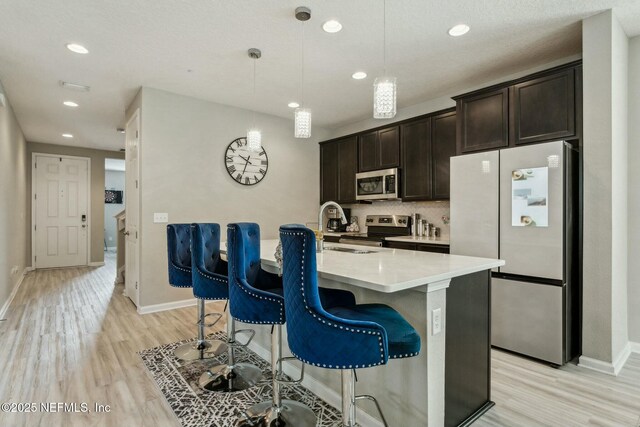 kitchen with dark brown cabinetry, a kitchen island with sink, stainless steel appliances, light wood-style floors, and light countertops