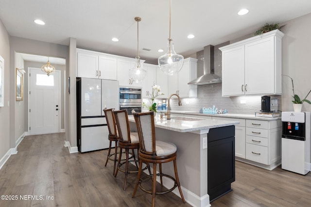 kitchen with white cabinets, a center island with sink, white fridge, and wall chimney range hood