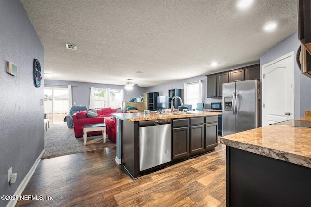 kitchen featuring dark wood-type flooring, a textured ceiling, appliances with stainless steel finishes, an island with sink, and ceiling fan