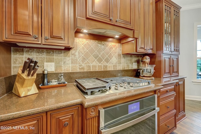 kitchen featuring crown molding, backsplash, stainless steel appliances, custom range hood, and light wood-type flooring