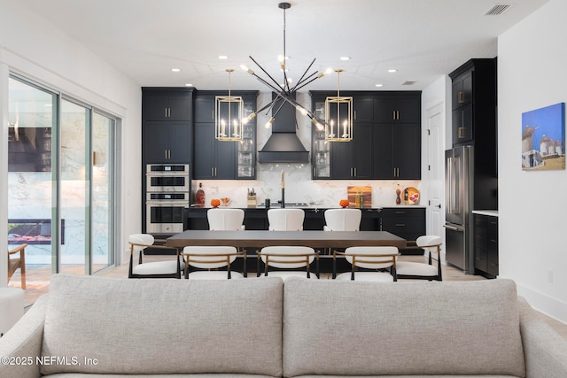 kitchen with premium range hood, a chandelier, hanging light fixtures, stainless steel appliances, and a center island with sink