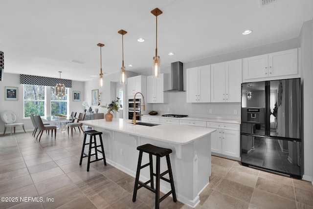 kitchen featuring wall chimney exhaust hood, white cabinetry, an island with sink, pendant lighting, and black appliances