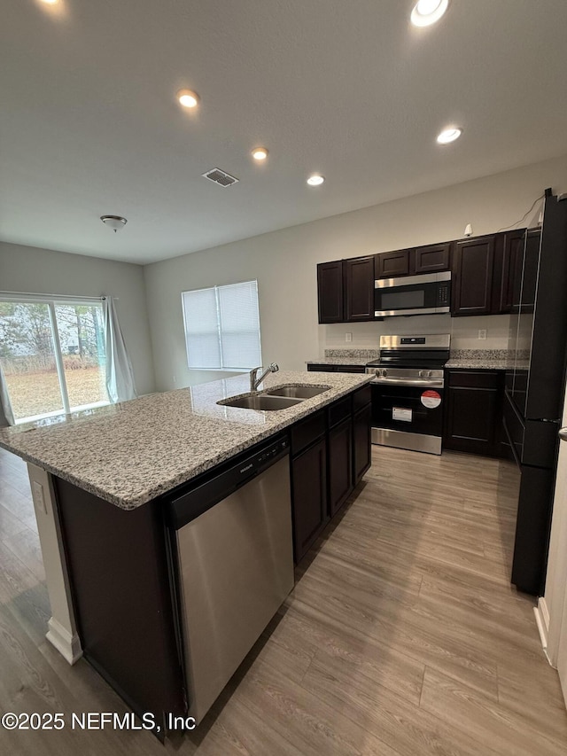 kitchen with sink, stainless steel appliances, light stone countertops, an island with sink, and light wood-type flooring