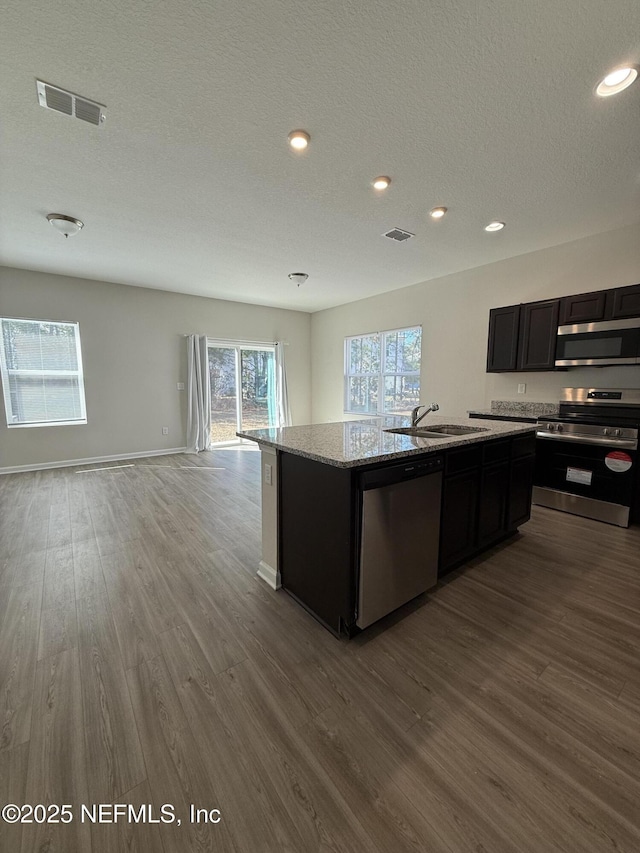 kitchen with sink, a center island with sink, appliances with stainless steel finishes, hardwood / wood-style flooring, and light stone countertops