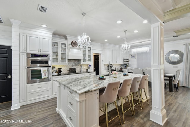 kitchen featuring double oven, decorative light fixtures, white cabinetry, light stone countertops, and a center island with sink