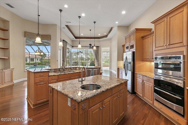 kitchen featuring brown cabinetry, a center island with sink, a tray ceiling, and a sink