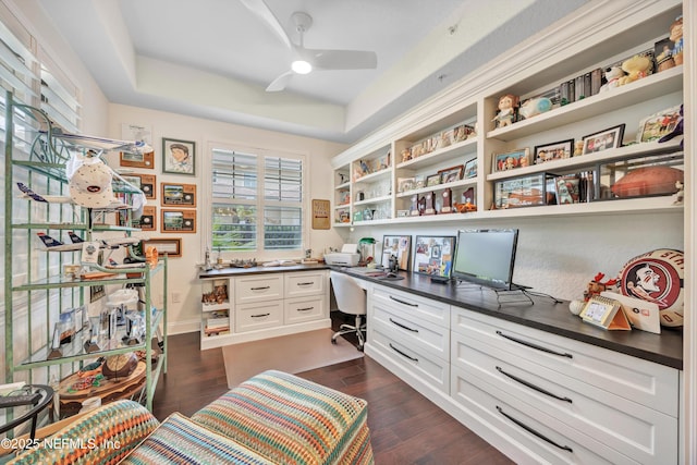 home office with dark wood-type flooring, ceiling fan, built in desk, and a raised ceiling