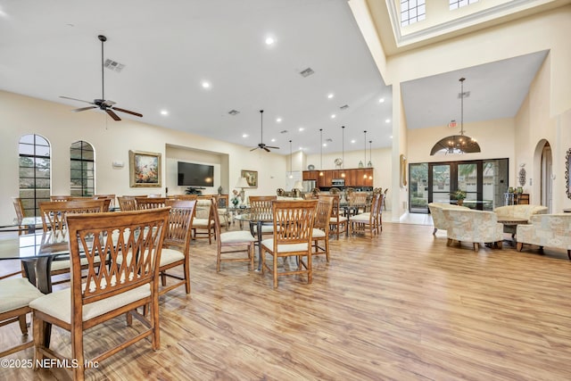 dining space featuring a towering ceiling, plenty of natural light, ceiling fan with notable chandelier, and light wood-type flooring