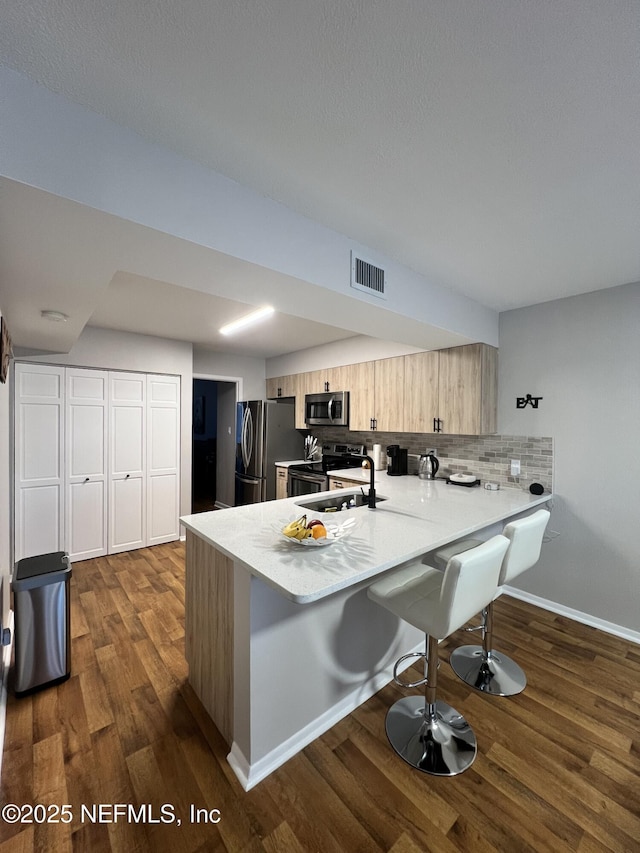 kitchen with appliances with stainless steel finishes, a breakfast bar, light brown cabinetry, dark hardwood / wood-style flooring, and kitchen peninsula