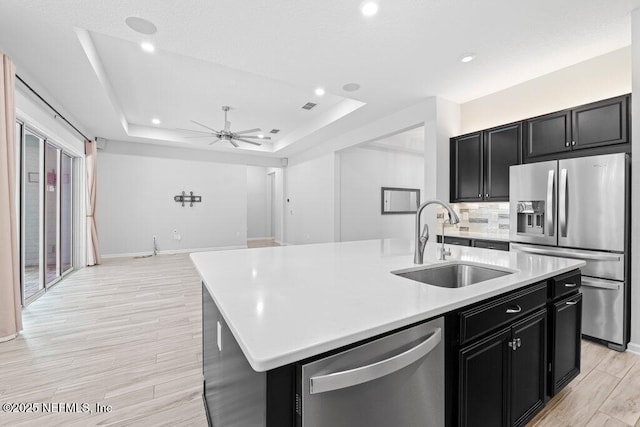 kitchen featuring sink, ceiling fan, appliances with stainless steel finishes, a kitchen island with sink, and a tray ceiling