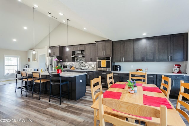 kitchen with decorative light fixtures, backsplash, exhaust hood, stainless steel appliances, and a center island with sink