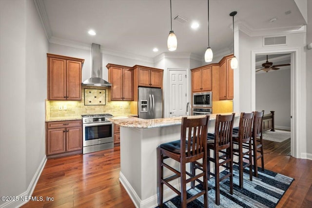 kitchen featuring a kitchen bar, light stone counters, decorative light fixtures, stainless steel appliances, and wall chimney range hood