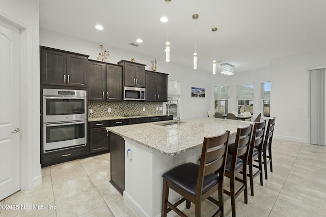 kitchen featuring a breakfast bar area, a kitchen island with sink, stainless steel appliances, light stone countertops, and decorative light fixtures