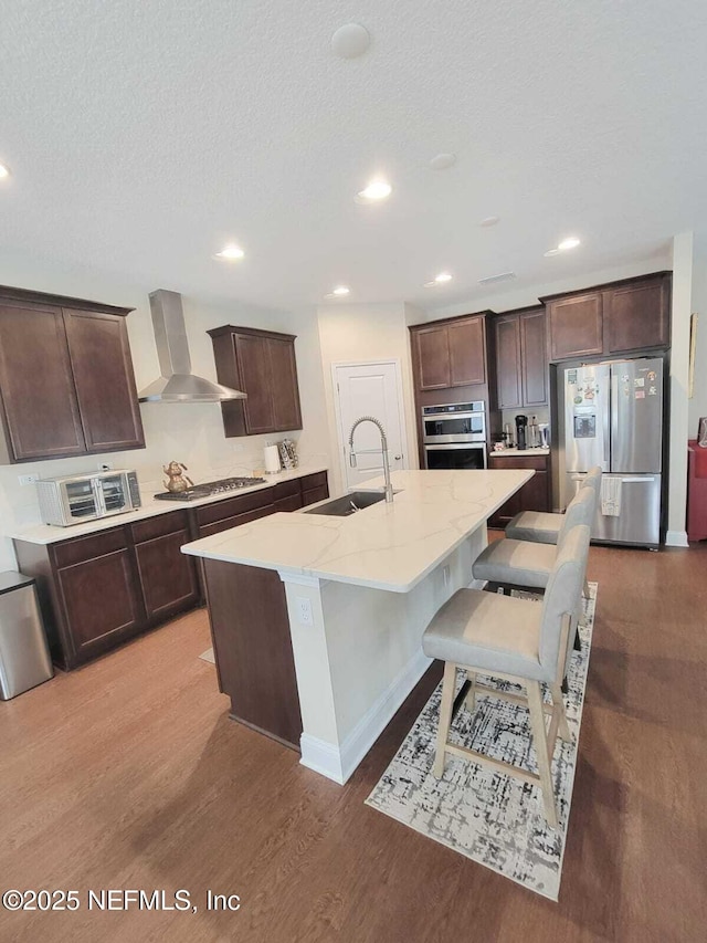 kitchen with sink, a breakfast bar area, a kitchen island with sink, stainless steel appliances, and wall chimney exhaust hood