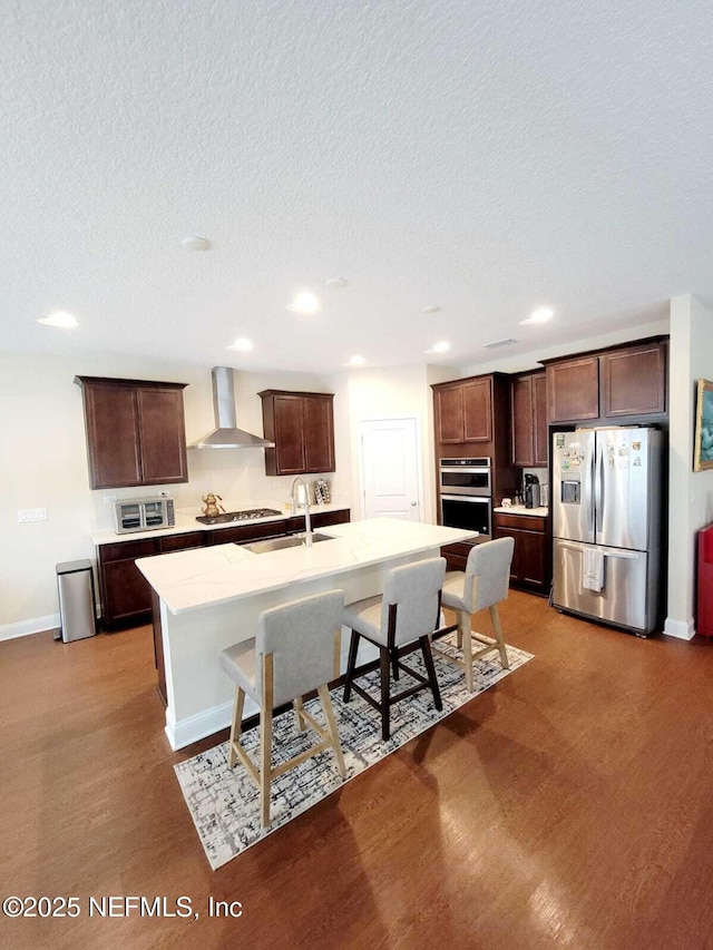 kitchen with sink, a breakfast bar area, a center island with sink, stainless steel appliances, and wall chimney range hood