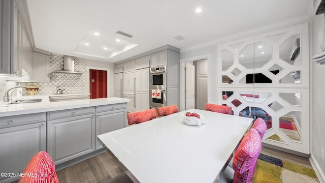 kitchen with gray cabinets, double oven, sink, decorative backsplash, and wall chimney range hood