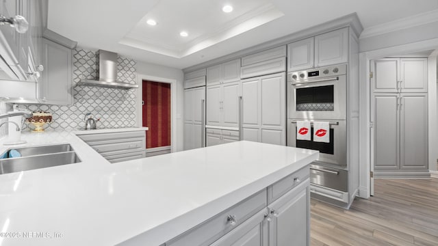 kitchen with sink, gray cabinetry, a tray ceiling, double oven, and wall chimney range hood