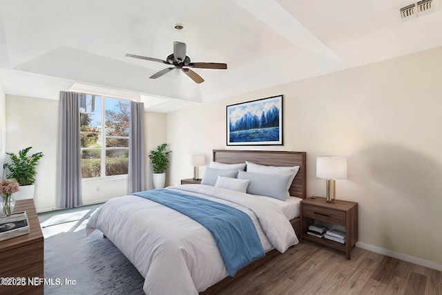 bedroom featuring wood finished floors, a ceiling fan, visible vents, baseboards, and a tray ceiling