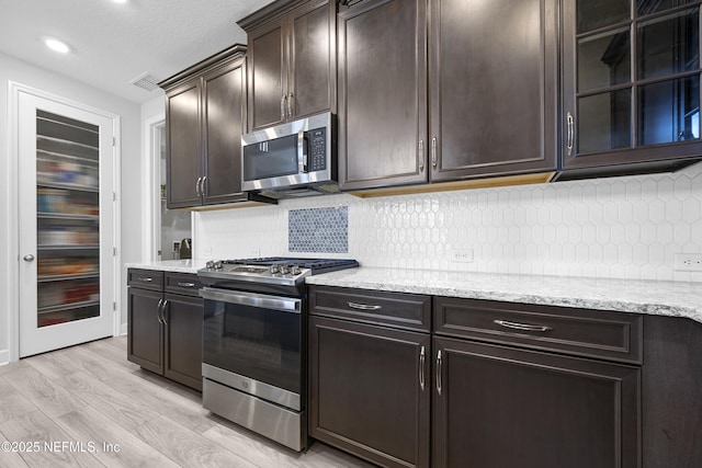 kitchen with tasteful backsplash, light stone counters, dark brown cabinets, light wood-type flooring, and appliances with stainless steel finishes