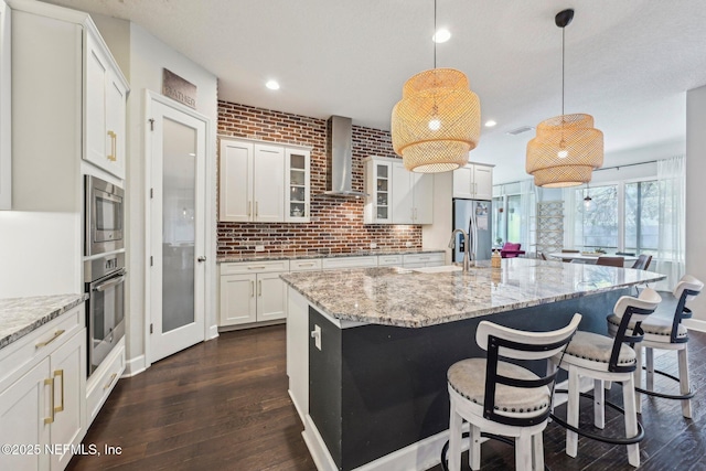 kitchen with wall chimney exhaust hood, decorative light fixtures, appliances with stainless steel finishes, an island with sink, and white cabinets