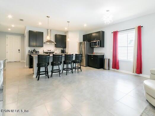 kitchen featuring wall chimney exhaust hood, a breakfast bar area, hanging light fixtures, appliances with stainless steel finishes, and an island with sink