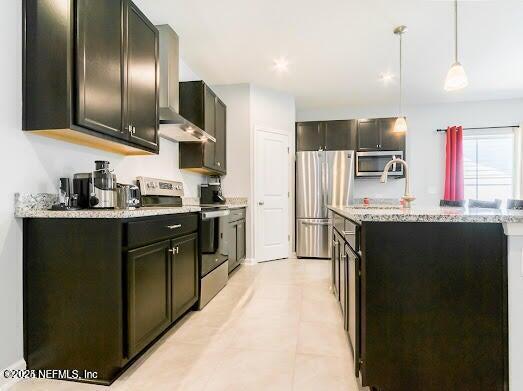 kitchen with wall chimney exhaust hood, decorative light fixtures, appliances with stainless steel finishes, an island with sink, and light stone countertops