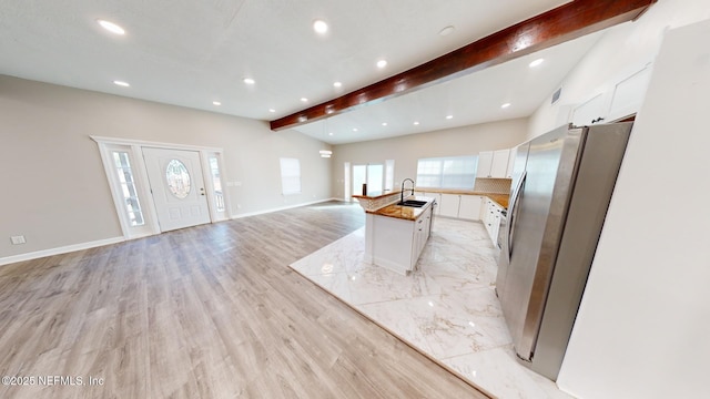 kitchen with stainless steel refrigerator, lofted ceiling with beams, sink, white cabinets, and a center island with sink