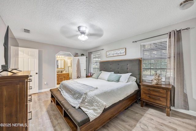 bedroom with a textured ceiling, ensuite bath, light hardwood / wood-style floors, and ceiling fan