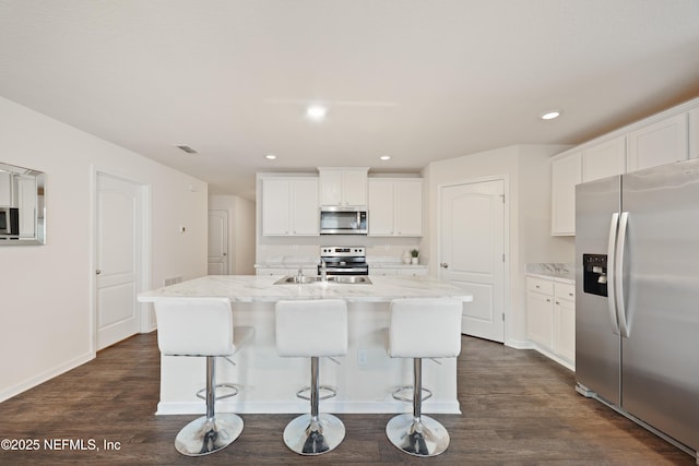 kitchen featuring appliances with stainless steel finishes, an island with sink, white cabinets, light stone countertops, and dark wood-type flooring