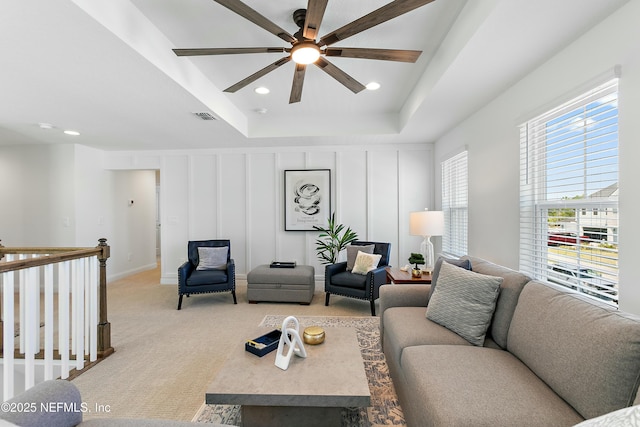 living area with visible vents, a raised ceiling, a decorative wall, a ceiling fan, and light carpet