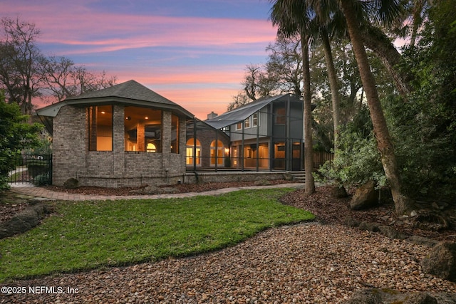 back of house at dusk with a sunroom, brick siding, a lawn, and fence