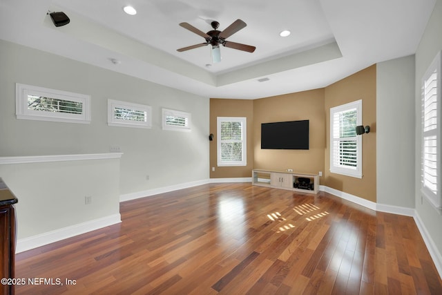 unfurnished living room with a ceiling fan, wood finished floors, baseboards, a healthy amount of sunlight, and a raised ceiling