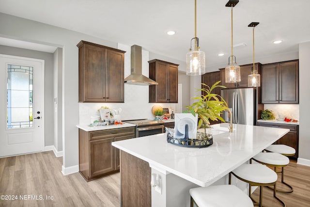 kitchen featuring tasteful backsplash, decorative light fixtures, appliances with stainless steel finishes, an island with sink, and wall chimney range hood