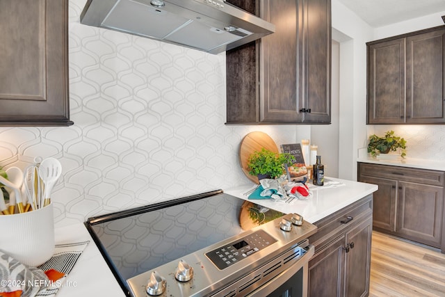 kitchen featuring electric stove, tasteful backsplash, dark brown cabinets, wall chimney range hood, and light hardwood / wood-style flooring