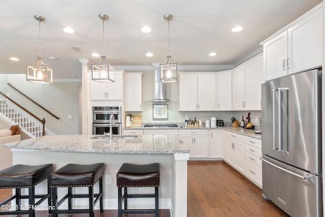 kitchen with wall chimney range hood, appliances with stainless steel finishes, white cabinets, a center island with sink, and decorative light fixtures