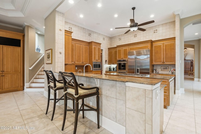 kitchen with sink, built in appliances, a kitchen breakfast bar, light stone countertops, and a kitchen island with sink